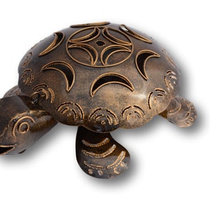 May include: A bronze-coloured metal turtle figurine with a dark, weathered finish. The shell features cut-out patterns including crescent moons and circular motifs. The turtle has a head, four legs, and a tail, with etched detailing on its limbs.