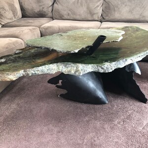 May include: A unique coffee table with a green stone top and a black metal base. The stone top is shaped like a jagged rock and has a rough texture. The base is made of two pieces of metal that are shaped like abstract forms. The table is a modern and stylish piece of furniture that would be a great addition to any home.