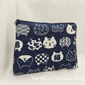 Könnte beinhalten: Eine marineblaue Reißverschlusstasche mit einem sich wiederholenden Muster aus weißen Katzenköpfen und abstrakten Designs. Die Tasche hat eine rechteckige Form und einen dunkelblauen Reißverschluss. Der Text "Herzlut Designs.etsy.com" ist unten sichtbar.