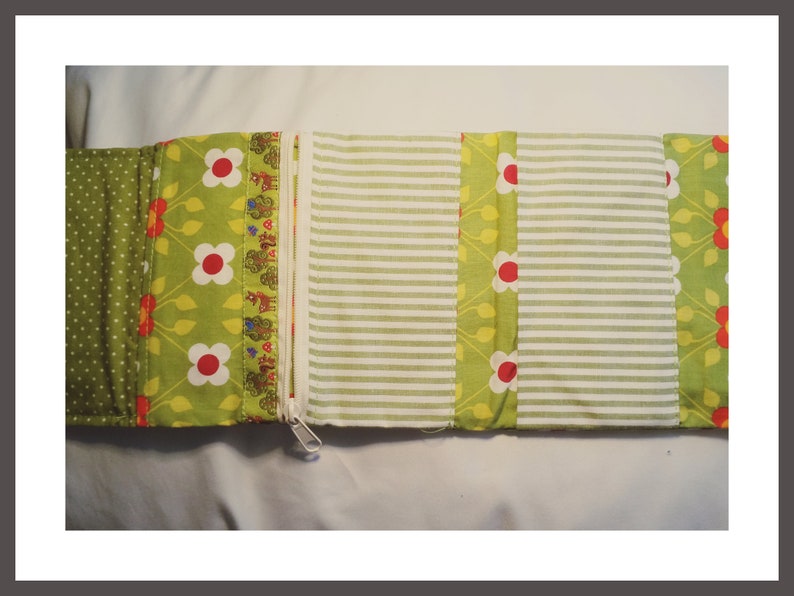 K&ouml;nnte beinhalten: Ein Stoff-Organizer mit Rei&szlig;verschluss, der verschiedene gemusterte Abschnitte aufweist. Das Design umfasst gr&uuml;ne Punkte, Blumendrucke mit roten und wei&szlig;en Blumen sowie wei&szlig;-gr&uuml;ne Streifen. Der Organizer ist offen und flach.