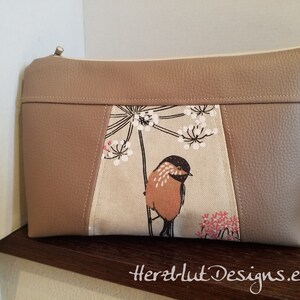 Könnte beinhalten: Beige Clutch-Tasche mit Vogel- und Blumenmuster. Sie hat einen beigen Reißverschluss und einen Riemen. Das Mittelteil zeigt einen Vogel auf einem Ast mit weißen Blüten und rosa Akzenten. Der Text "Herzlut Designs.etsy" ist sichtbar.