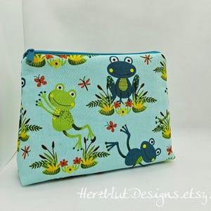 Könnte beinhalten: Hellblaue Reißverschlusstasche mit einem skurrilen Froschmuster. Das Design umfasst grüne und blaue Frösche, rote Schmetterlinge und florale Akzente. Der Text "Herztlut Designs.etsy.com" ist unten aufgedruckt.