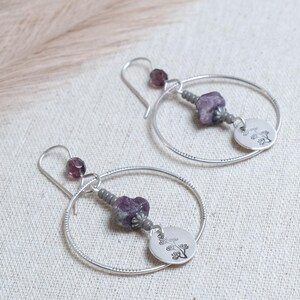 May include: Silver hoop earrings with purple and gray beads. Each earring features a small, round charm with a tree design. The earrings have a simple, elegant design, perfect for everyday wear. The earrings are displayed on a neutral-colored fabric.