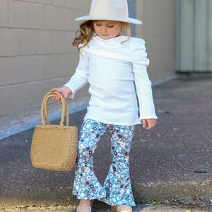 May include: A young girl wearing a white sweater, blue and white floral bell bottom pants, a white hat, and holding a brown woven tote bag.