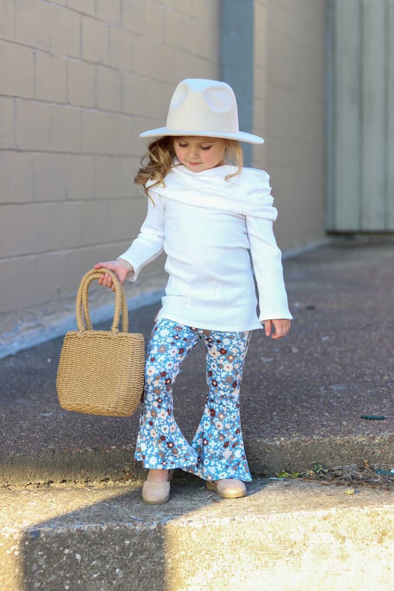 May include: A young girl wearing a white sweater, blue and white floral bell bottom pants, a white hat, and holding a brown woven tote bag.