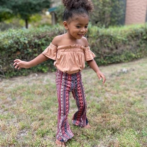 May include: A young girl wearing a peach off-the-shoulder top and bell bottom pants with a colorful geometric pattern.