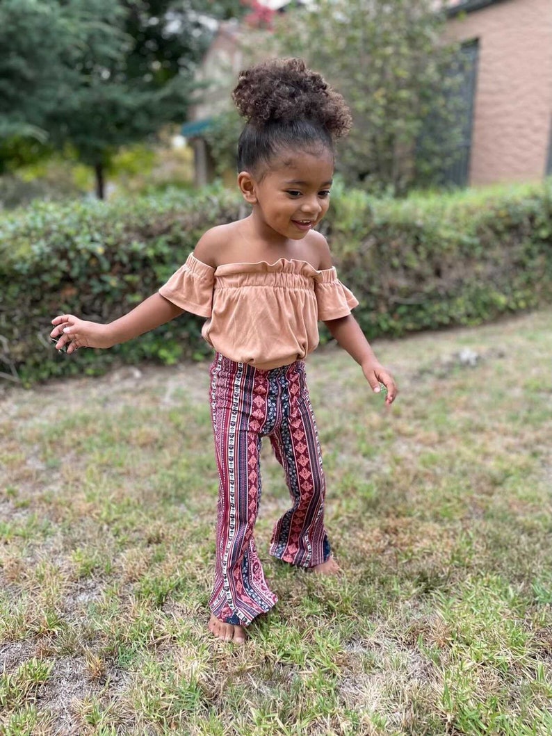May include: A young girl wearing a peach off-the-shoulder top and bell bottom pants with a colorful geometric pattern.