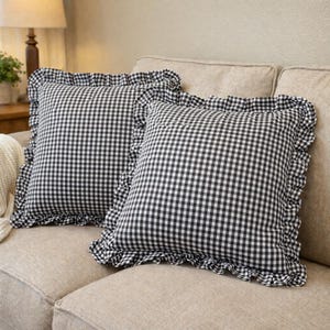 May include: Two square throw pillows with a black and white gingham pattern and ruffled edges. The pillows are on a beige sofa, creating a cozy and classic aesthetic. The pillows add a touch of farmhouse charm to any living space.