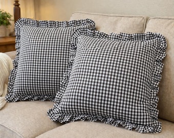 May include: Two square throw pillows with a black and white gingham pattern and ruffled edges. The pillows are on a beige sofa, creating a cozy and classic aesthetic. The pillows add a touch of farmhouse charm to any living space.