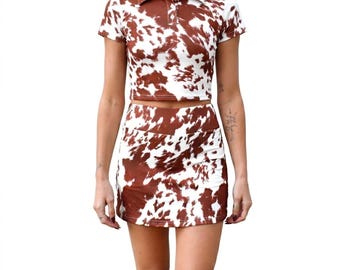 May include: A two-piece set consisting of a short-sleeved polo crop top and a matching mini skirt. Both pieces feature a brown and white cow print pattern. The polo top has a collar and a button-down placket with visible white buttons.