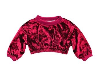 May include: A cropped, long-sleeved top in a deep red velvet fabric. The top features a crew neck and elasticated hem and cuffs. The fabric has a textured appearance, with a slight sheen. The sleeves are a three-quarter length.