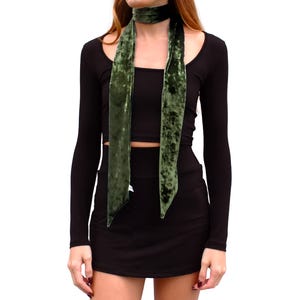 May include: A long, olive green velvet scarf tied around the neck, featuring a soft, crushed texture and pointed ends. The scarf is worn over a black long-sleeved crop top and a matching black mini skirt, creating a simple, layered ensemble.