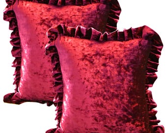 May include: Two square, plush throw pillows in a deep red-burgundy hue. Each pillow features a ruffled edge, adding a decorative touch. The velvet-like texture of the pillows creates a luxurious and inviting aesthetic, perfect for home decor.