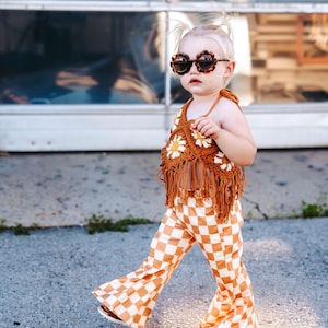 May include: A young child wearing a brown crocheted top with white daisy accents and brown fringe. The child is also wearing brown and white checkered bell bottom pants and tortoise shell sunglasses.