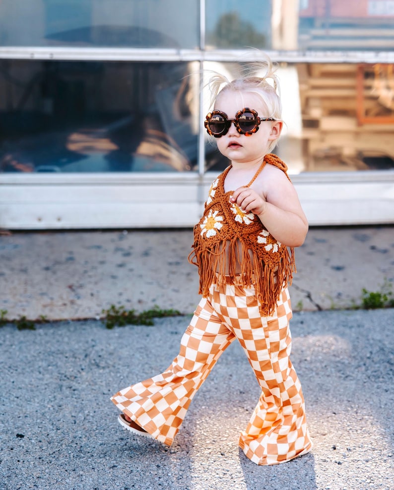 May include: A young child wearing a brown crocheted top with white daisy accents and brown fringe. The child is also wearing brown and white checkered bell bottom pants and tortoise shell sunglasses.