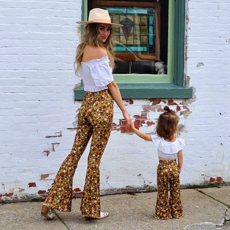 May include: A woman and a young girl wearing matching floral print bell bottom pants. The woman is wearing a white off-the-shoulder top and a straw hat. The girl is wearing a white ruffled top.