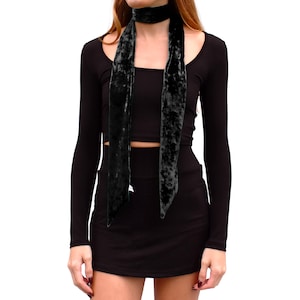 May include: A long, narrow black velvet scarf with a crushed texture, worn wrapped around the neck. The scarf features two long, pointed ends that hang down over the chest. It is paired with a black long-sleeved crop top and a matching black skirt.
