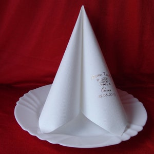Könnte beinhalten: Eine weiße Papierserviette, die zu einem Kegel gefaltet ist und auf einem weißen Teller liegt. Die Serviette hat eine goldene Aufschrift mit dem Text "Meine Taufe", einem Schneeflocke und einem kleinen Bild eines Kinderwagens. Der Text enthält auch den Namen "Clemens" und das Datum "22.06.2013".