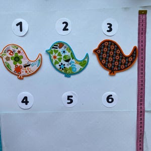 Könnte beinhalten: Drei farbenfrohe Vogel-Applikationen aus Stoff. Die Vögel haben orange, blaue und grüne Umrisse mit floralen und geometrischen Mustern in verschiedenen Farben. Ein Maßband ist auf der rechten Seite sichtbar.
