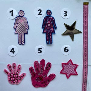 Könnte beinhalten: Eine Sammlung von Stoffapplikationen in verschiedenen Formen. Enthält zwei weibliche Figuren, einen Stern und zwei Handformen, alle mit Blumen- oder Karomustern. Ein rosa Maßband ist auf der rechten Seite sichtbar.