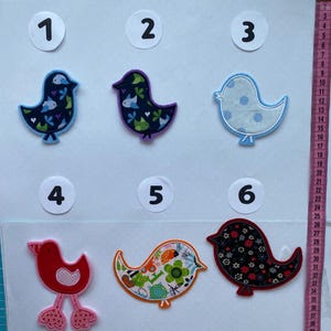 Könnte beinhalten: Sechs bunte Vogel-förmige Stoffaufnäher. Jeder Aufnäher hat ein einzigartiges Design, darunter Blumenmuster, Tupfen und unifarbene Farben. Die Aufnäher sind mit 1 bis 6 nummeriert und eignen sich ideal für Näh- oder Bastelprojekte.