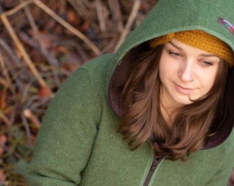 Puede incluir: Una mujer con una chaqueta verde con capucha y forro marrón y una diadema amarilla. Está sosteniendo una pequeña rama de pino en sus manos.