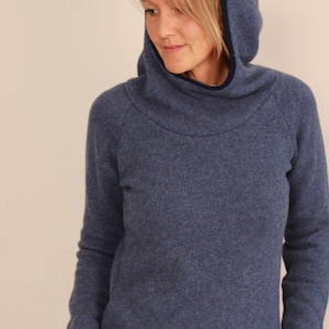 May include: A blue hooded sweater with a high neckline and a dark blue trim around the hood. The sweater has a ribbed texture and long sleeves. The model is wearing the sweater.