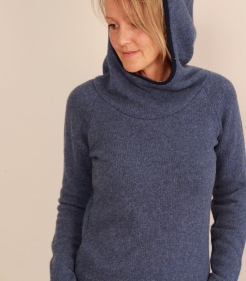 May include: A blue hooded sweater with a high neckline and a dark blue trim around the hood. The sweater has a ribbed texture and long sleeves. The model is wearing the sweater.