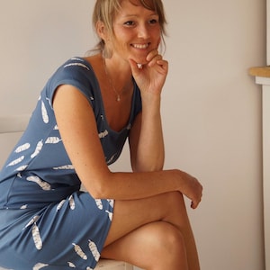 May include: A woman wearing a blue dress with a white feather pattern. She is sitting on a white chair and smiling.