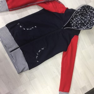 May include: A navy blue hooded sweatshirt with red sleeves and a white anchor print on the hood. The sweatshirt has a white and black striped band at the bottom and cuffs.