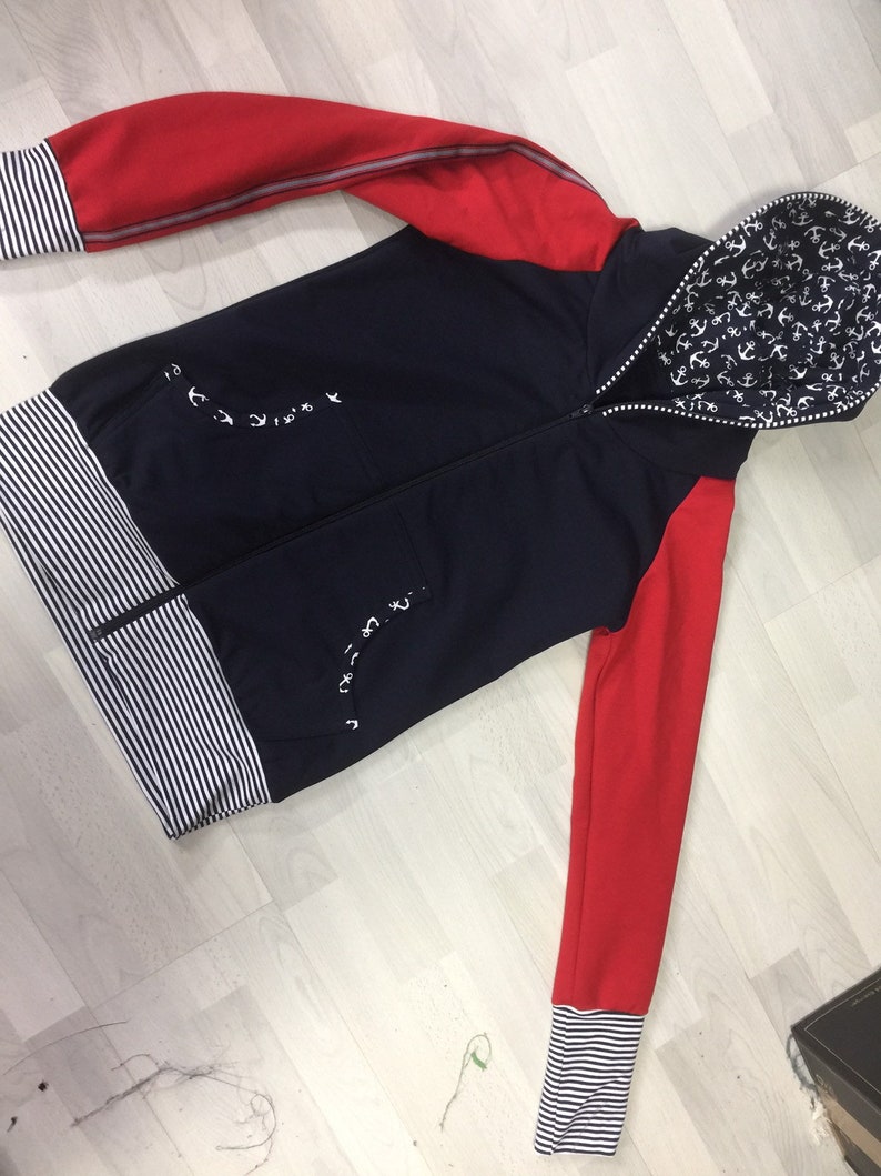 May include: A navy blue hooded sweatshirt with red sleeves and a white anchor print on the hood. The sweatshirt has a white and black striped band at the bottom and cuffs.