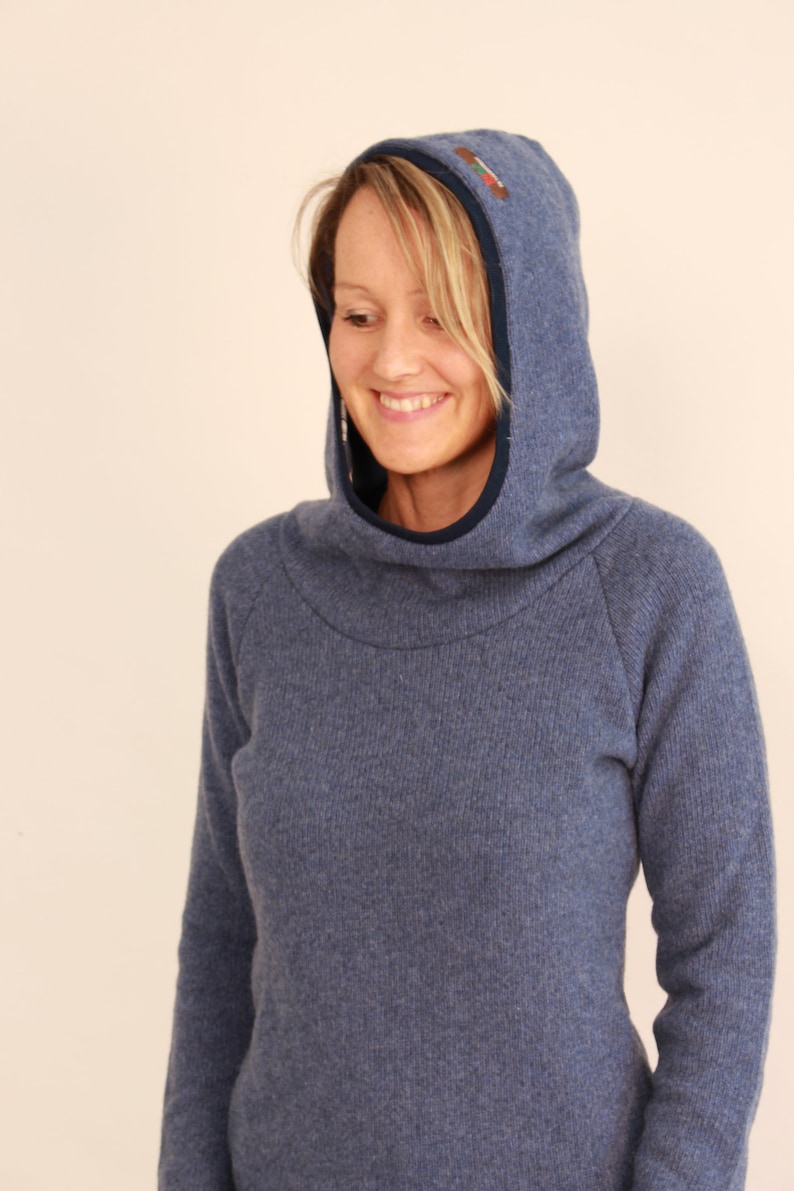 May include: A blue hooded sweater with a high collar and a small embroidered detail on the hood. The sweater has long sleeves and a comfortable, relaxed fit. The hood is lined with a darker blue fabric.