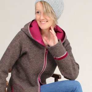 Könnte beinhalten: Eine braune Kapuzenjacke mit rosa Futter und Reißverschluss. Die Jacke hat einen weißen Besatz um die Kapuze und den Reißverschluss. Das Model trägt eine graue Strickmütze.