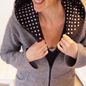 Könnte beinhalten: Eine graue Kapuzenjacke mit einem schwarzen und weißen Tupfenfutter. Die Jacke hat einen Reißverschluss und zwei Taschen. Die Jacke ist mit einem schwarzen und weißen karierten Stoff gefüttert.