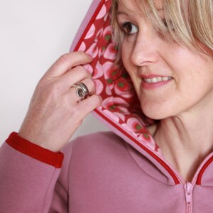 May include: A pink hooded sweatshirt with a red zip and red trim. The hood is lined with a floral fabric in shades of pink, red, and green.