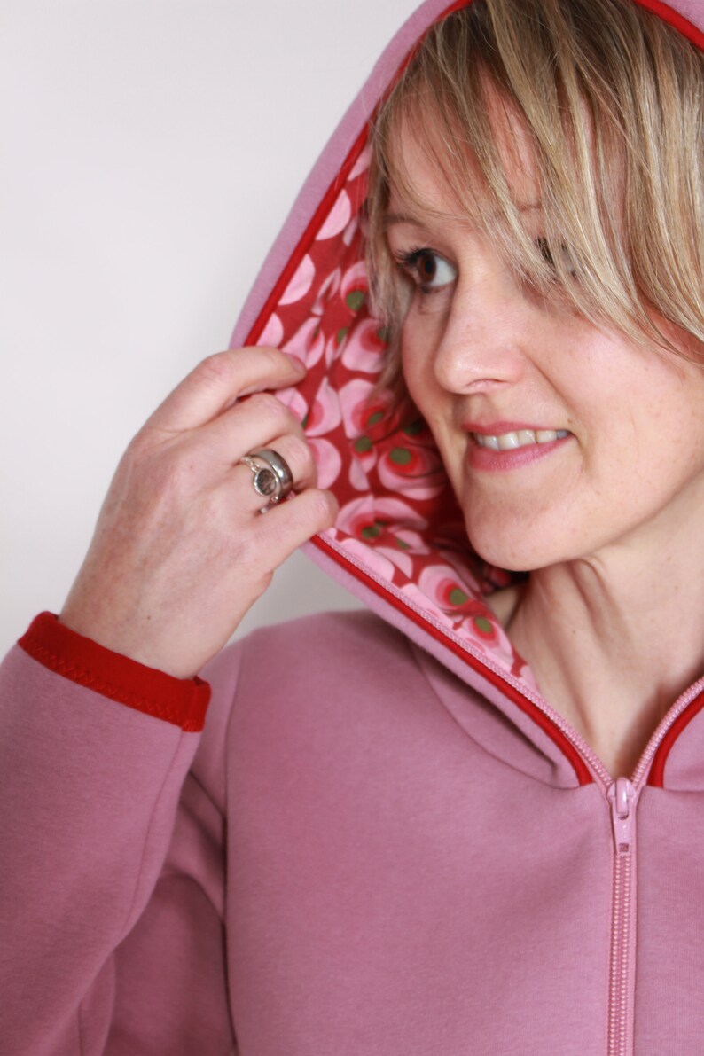 May include: A pink hooded sweatshirt with a red zip and red trim. The hood is lined with a floral fabric in shades of pink, red, and green.