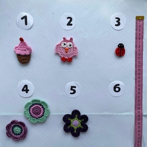 Könnte beinhalten: Eine Sammlung handgefertigter Häkelapplikationen, darunter ein rosa Cupcake, eine Eule, ein Marienkäfer und Blumen. Die Applikationen sind in verschiedenen Farben gehalten, mit einem Maßband in Zentimetern zur Größenbestimmung.