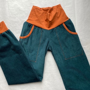 Könnte beinhalten: Tealfarbene Cordhose mit orangefarbenem Bund und Bündchen. Die Hose hat zwei Vordertaschen mit orangefarbenem Besatz und sichtbaren Nähten. Der Stoff hat eine vertikale Rippenstruktur.