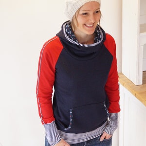 May include: A navy blue hooded sweatshirt with red sleeves and white trim. The sweatshirt has a cowl neck and a small pocket on the front. The model is wearing a white knit hat.
