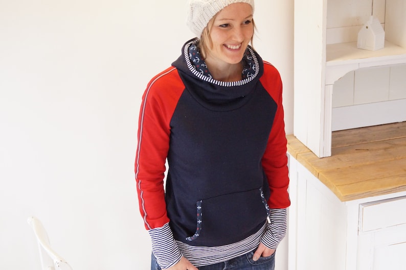 May include: A navy blue hooded sweatshirt with red sleeves and white trim. The sweatshirt has a cowl neck and a small pocket on the front. The model is wearing a white knit hat.