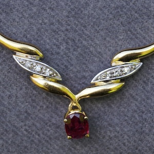 Könnte beinhalten: Eine goldene Halskette mit einem roten Edelstein-Anhänger und Diamant-Akzenten. Die Halskette hat ein zartes, geschwungenes Design.