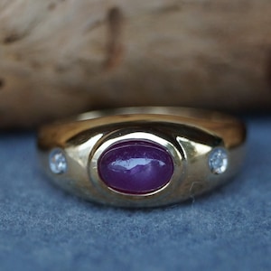 Könnte beinhalten: Ein goldener Ring mit einem großen ovalen Rubin in der Mitte und zwei kleineren, runden, klaren Edelsteinen. Der Ring ist vor einem neutralen Hintergrund abgebildet.