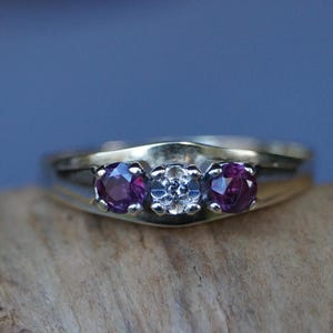 Könnte beinhalten: Ein Goldring mit drei runden Edelsteinen. Der mittlere Stein ist weiß und die beiden äußeren Steine sind lila.