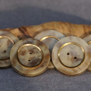 Könnte beinhalten: Sechs große, runde Knöpfe. Jeder Knopf hat ein hellbeige-braunes Marmormuster mit einem goldfarbenen Ring in der Mitte. Jeder Knopf hat zwei Löcher zum Annähen.