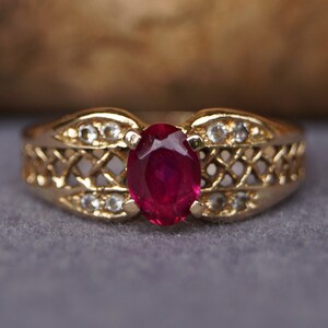 Könnte beinhalten: Ein goldener Ring mit einem ovalen, tiefroten Edelstein in der Mitte, flankiert von kleineren, klaren Steinen. Das Band hat ein durchbrochenes Gittermuster. Der Ring liegt auf einer grauen Oberfläche.