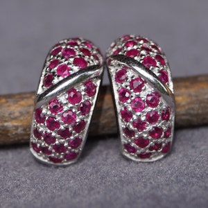 Könnte beinhalten: Ein Paar silberne Creolen mit einem Kreuzdesign und kleinen roten Rubin-Steinen.