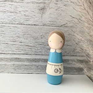 May include: A wooden doll with a light brown head, blue eyes, and pink cheeks. The doll is wearing a blue dress with a white apron and a floral design. The doll is standing on a white surface.