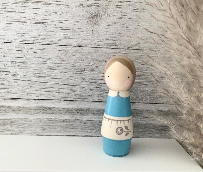 May include: A wooden doll with a light brown head, blue eyes, and pink cheeks. The doll is wearing a blue dress with a white apron and a floral design. The doll is standing on a white surface.