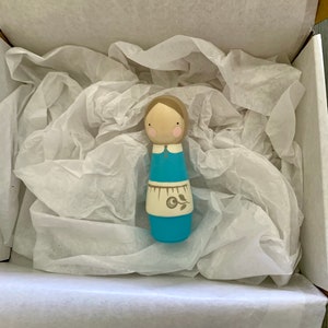 May include: A wooden doll with a blue dress, white apron, and brown hair. The doll is in a white tissue-filled box.