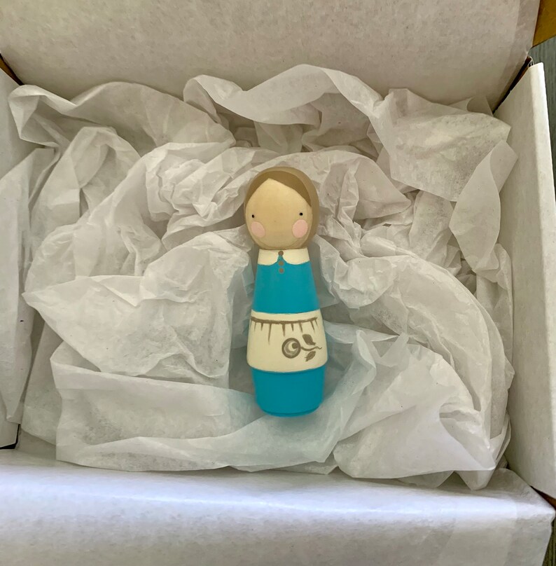 May include: A wooden doll with a blue dress, white apron, and brown hair. The doll is in a white tissue-filled box.